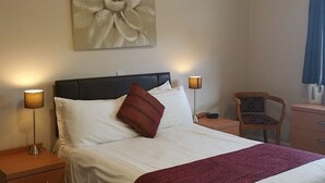 En snyggt bäddad säng med vita sängkläder och en röd pläd, med två sänglampor på varsin sida. Ett stort, blommigt konstverk hänger på väggen ovanför sängen.