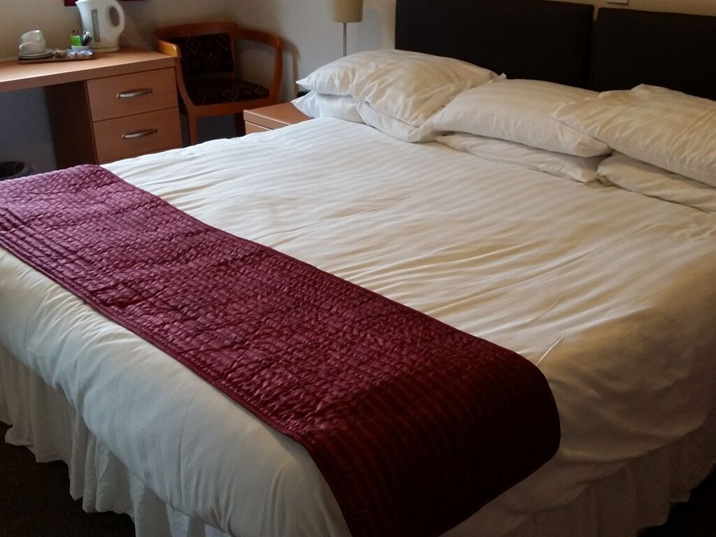 Una cama bien hecha con sábanas blancas y una manta roja, una mesita de noche de madera con una lámpara, y una silla con cojín.