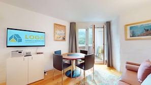 Ein modernes Wohnzimmer mit Fernseher und einem Logo, ein Essbereich mit rundem Tisch und Stühlen sowie ein Blick auf einen Balkon mit einer Tür.
