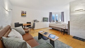 A modern, well-lit living room with a brown leather sofa, a dining area with a table and chairs, a bed with a view, and a TV on the wall. - Studio apartment on the 3rd floor with balcony, approx. (Champfèr)