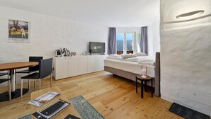 A modern hotel room with a wooden floor, a bed with a view, a desk with a chair, a TV, and a minibar. - Studio apartment on the 3rd floor with balcony, approx. (Champfèr)