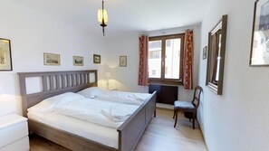 A neatly made bed with a wooden headboard, a bedside table with a lamp, a chair, and a window with curtains.