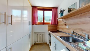 Eine kleine Küche mit weißen Schränken, einer Holzrückwand, einem Fenster mit roten Vorhängen und einer Spüle aus Edelstahl.