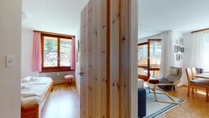 Ein Zimmer mit einem Bett, einem kleinen Hocker, einer Holztür und Blick auf ein Gebäude.