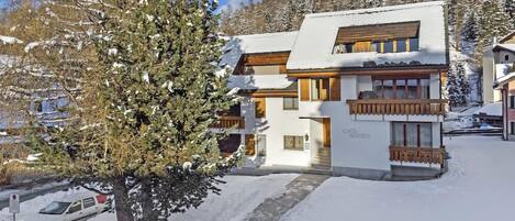 A two-story chalet with a wooden balcony, surrounded by snow-covered trees and a mountainous backdrop.