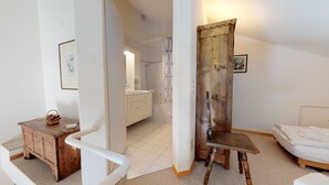A bedroom with a bed, a wooden chair, a small table with a basket, and a bathroom visible through an open door. - Spacious and quiet 2-room apartment on two floors, 45m2, facing north. (Sils/Segl Maria)