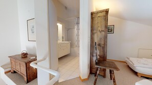 Une chambre à coucher avec un lit, une chaise en bois, une petite table avec un panier, et une salle de bain visible à travers une porte ouverte.