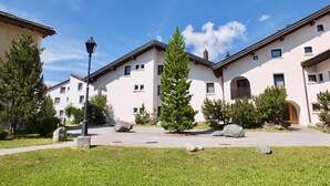 A row of modern buildings with a central courtyard, stone benches, and a street lamp. - Spacious and quiet 2-room apartment on two floors, 45m2, facing north. (Sils/Segl Maria)
