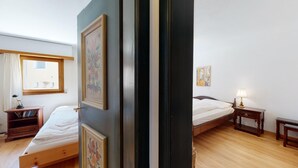 Ein Schlafzimmer mit zwei Betten, Holzboden, einem Fenster mit Vorhängen, einem Nachttisch mit Lampe und einem Bücherregal.