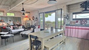 Un interior acogedor y rústico, con suelo de madera, una zona de comedor con banco y mesa, un conjunto de sofás, y un ventanal que ofrece vistas al exterior.