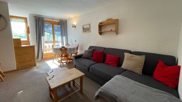 Ein gemütliches Wohnzimmer mit einem dunkelgrauen Sofa, einem Holztisch und einem Esstisch mit Stühlen. Ein Fenster mit Vorhängen, eine Holzkommode und ein Wandregal sind ebenfalls vorhanden.
