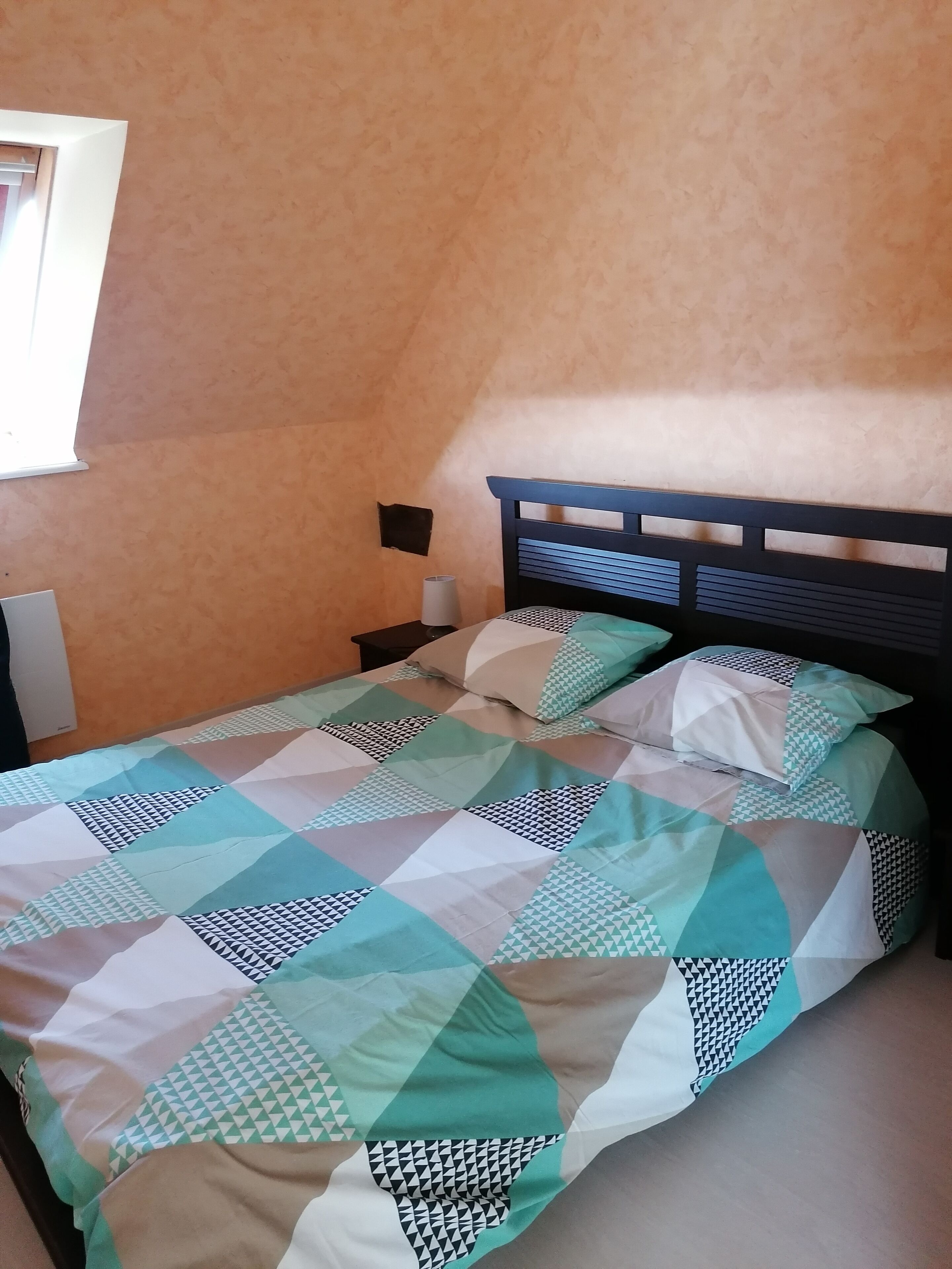 A bed with a geometric patterned bedspread, a wooden headboard, and a wall-mounted nightstand with a lamp.