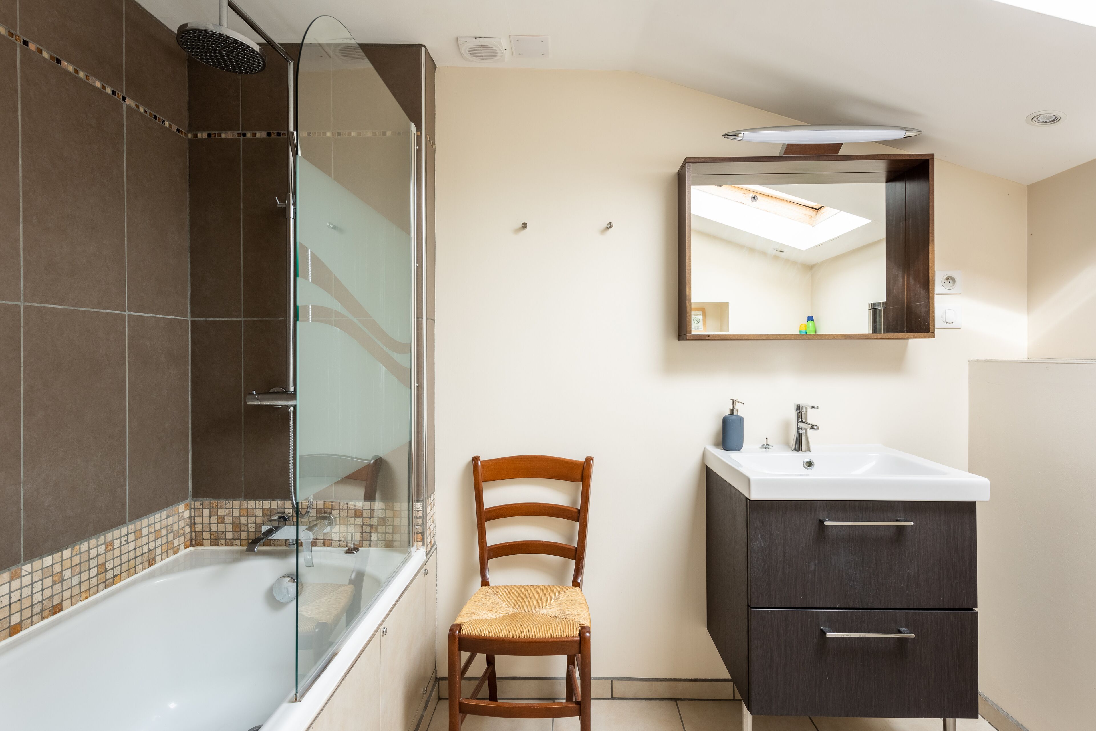 Un baño con ducha de vidrio, una silla de madera, un lavamanos con espejo y armarios oscuros.