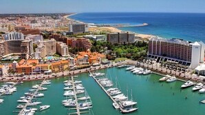 Una marina con numerose imbarcazioni ormeggiate, circondata da edifici e una spiaggia sullo sfondo.