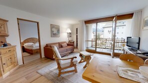 A cozy living room with a sofa, wooden chairs, a table, a TV, and a balcony with a view of buildings.