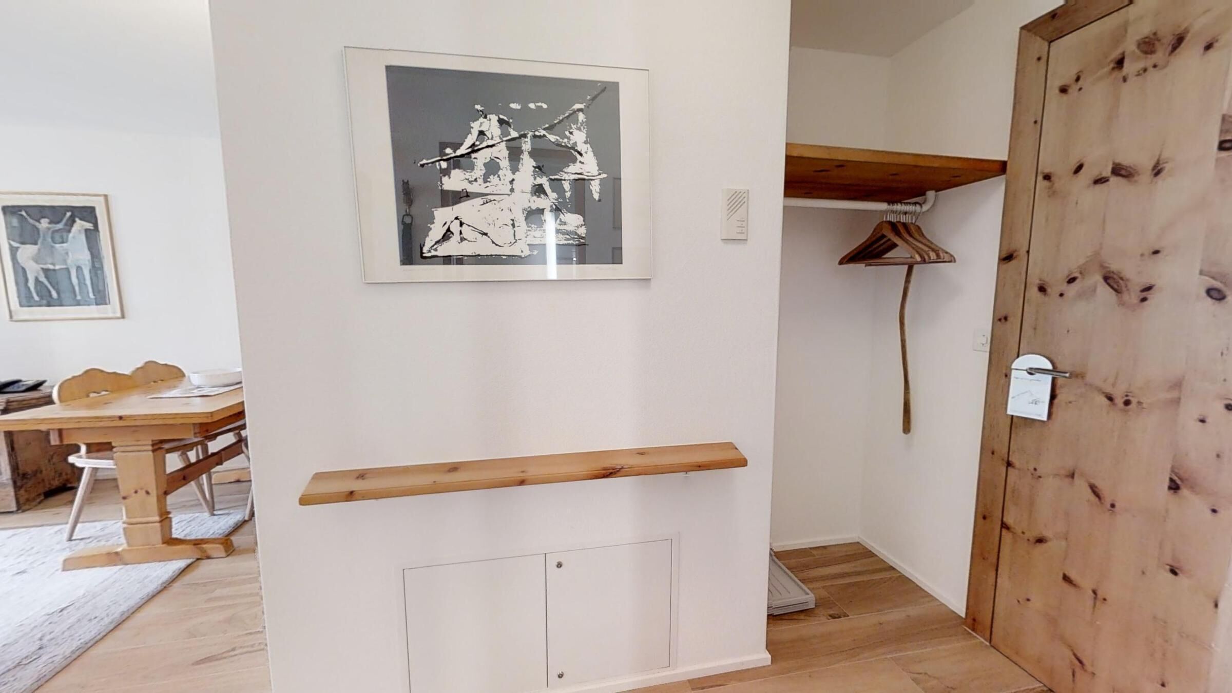 Una stanza con pavimento in legno, una parete bianca con un quadro incorniciato, una porta in legno e una piccola mensola di legno.
