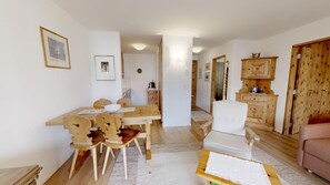 A dining area with a wooden table and chairs, a white armchair, a sofa, and a kitchen area with wooden cabinets and a wooden door.