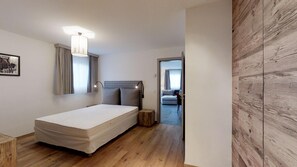 A modern bedroom with a wooden floor, a bed, a bedside table, a lamp, and a view into another room.