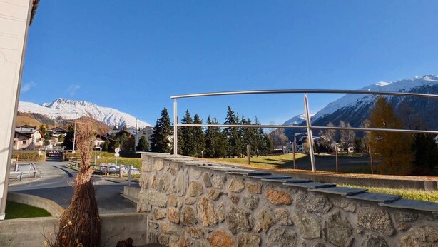 A stone wall with a metal railing overlooking a town with snow-capped mountains in the background. - 3-room apartment on the ground floor, about 90 m2. (Celerina/Schlarigna)