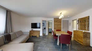 A modern living room with a sofa, a wooden dining table with chairs, a TV mounted on the wall, and a wooden accent wall.