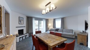 A modern living room with a wooden dining table, red chairs, a grey sofa, and a fireplace. - 3-room apartment on the ground floor, about 90 m2. (Celerina/Schlarigna)