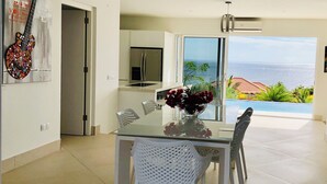 Eine moderne Küche mit Kühlschrank, einem Esstisch mit Stühlen und Blick auf den Ozean durch eine Glastür.