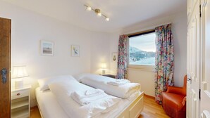 A double bed with white linens, a nightstand with a lamp, and a window with floral curtains.