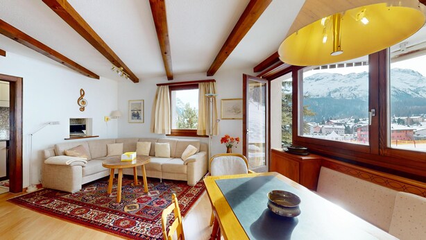 A cozy living room with a sofa, coffee table, and dining table. There is a large window with a view of a snowy mountain.