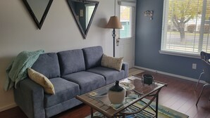 Un salon avec un canapé gris, une table basse et une grande fenêtre équipée de stores.