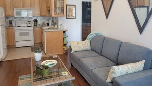 Une cuisine compacte avec des meubles en bois, un four à micro-ondes et une cuisinière. On y trouve une table basse en verre agrémentée d’objets décoratifs et un canapé gris avec des coussins à motifs.