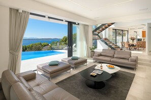 Uma sala de estar moderna com uma grande janela com vista para uma piscina e o mar, uma escadaria e um espaço de jantar.