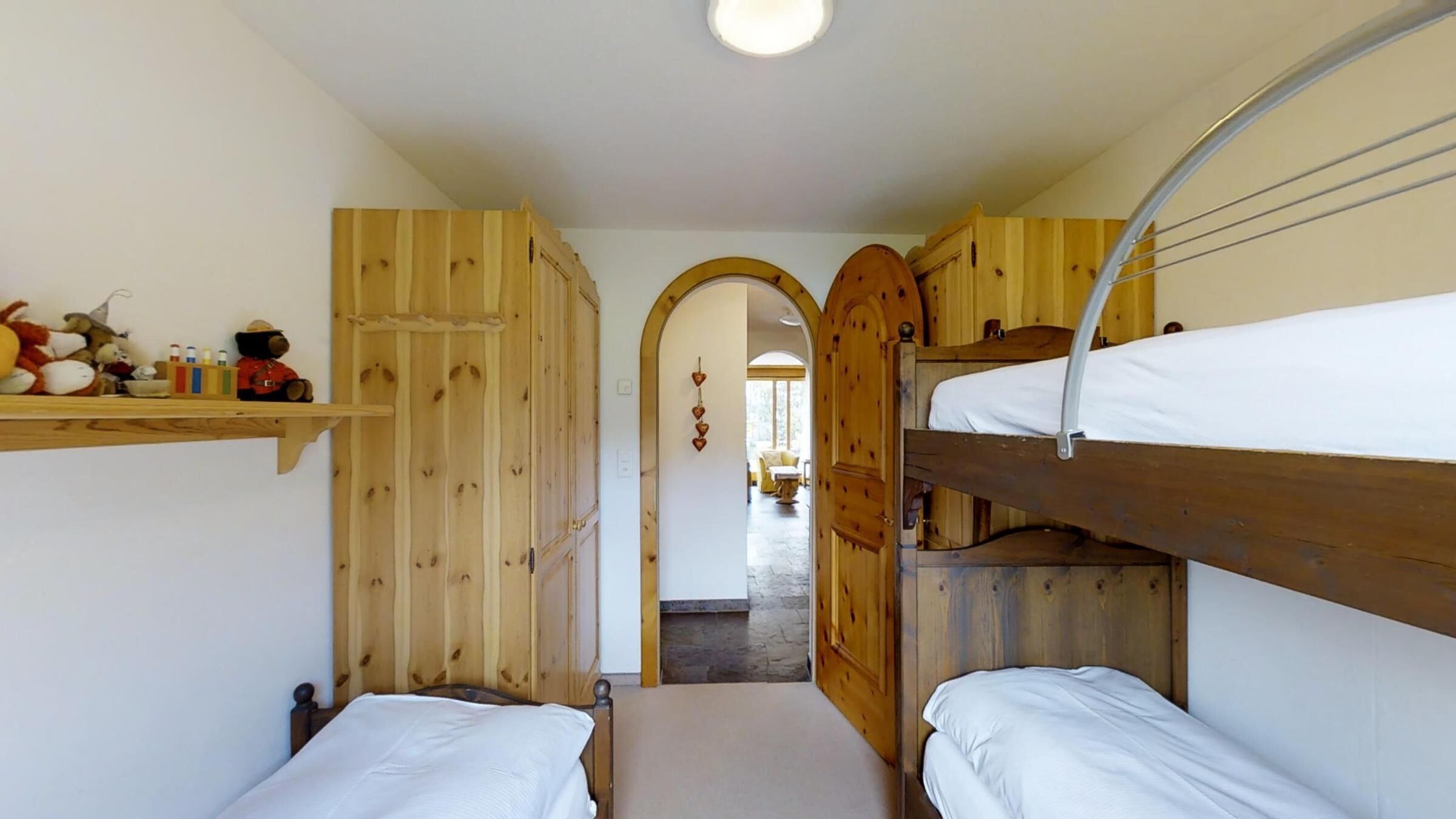 A narrow hallway with wooden bunk beds on either side, a shelf with toys, and a door leading to another room.