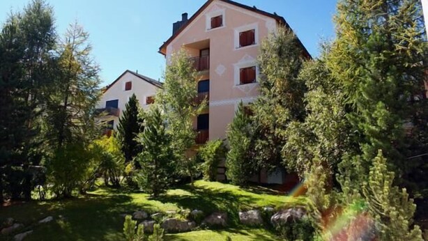 Un immeuble à plusieurs étages, doté d’un toit plat, entouré d’arbres et bordé d’une zone herbeuse parsemée de rochers.