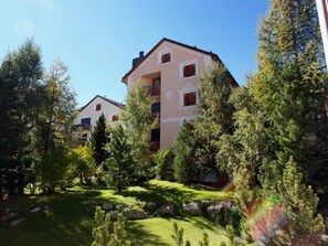 A multi-story building with a flat roof, surrounded by trees and a grassy area with rocks. - 3 - room apartment, about 70 m², on the ground floor. (Celerina/Schlarigna)