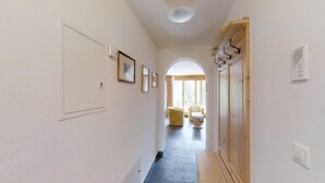Un couloir avec un banc en bois, une porte avec un porte-manteau, et une vue sur une pièce contenant un canapé et une table.