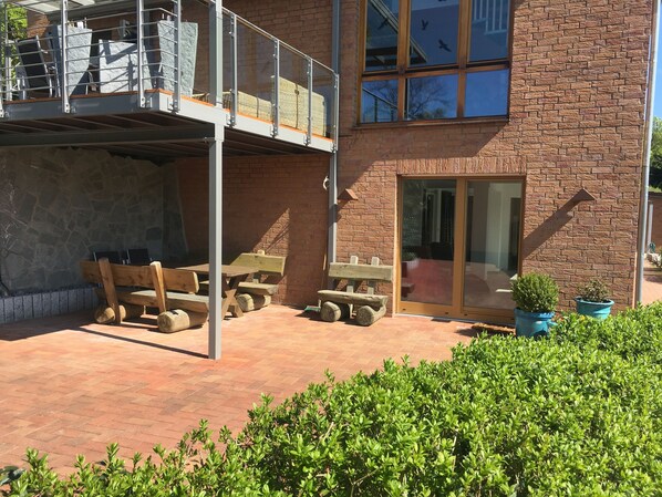 A building with a brick facade, a balcony with metal railings, and a patio area with wooden furniture and potted plants.