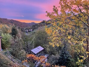 Un paisaje rural con una casa pequeña rodeada de follaje otoñal y colinas al fondo.