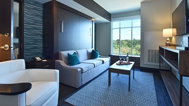 Una habitación de hotel moderna con sofá, una mesita con una bandeja de objetos, televisor y un amplio ventanal con vistas al exterior.