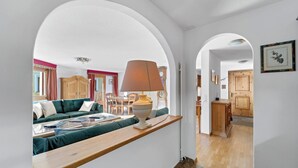 A spacious living area with a sofa, a wooden coffee table, and a lamp. There is a dining area with a table and chairs. The room has white walls and a wooden floor.