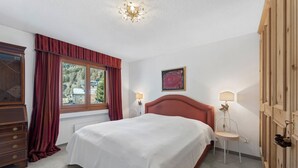 A bedroom with a bed, a dresser, a window with curtains, a painting, and a nightstand with a lamp.
