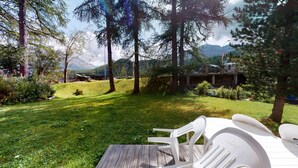 A wooden deck with white plastic chairs, surrounded by a grassy area with trees and a mountain view. - 5½-roomed apartment on the ground floor, 140m2. (Celerina/Schlarigna)