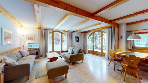 A spacious living room with a sofa, a coffee table, and a dining area with chairs. There is a television and a framed picture on the wall. - 5½-roomed apartment on the ground floor, 140m2. (Celerina/Schlarigna)