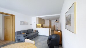 A modern living room with a grey sofa, a wooden coffee table, and a black armchair. A kitchen is visible in the background with a refrigerator and a microwave.