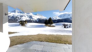 A view from a covered outdoor area with a stone floor, snow-covered landscape, and distant mountains. - 3-room apartment on the ground floor. (Silvaplana)