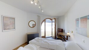 A bedroom with a bed, a desk, a chair, a round mirror, and a view of the beach through a door. - 3-room apartment on the ground floor. (Silvaplana)