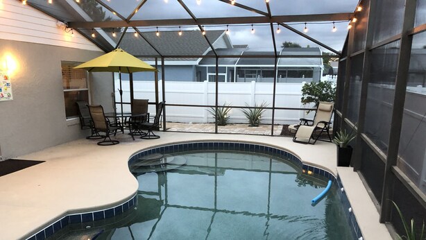 Ein Pool mit gewölbter Kante, umgeben von einer überdachten Terrasse mit Stühlen und Sonnenschirm.