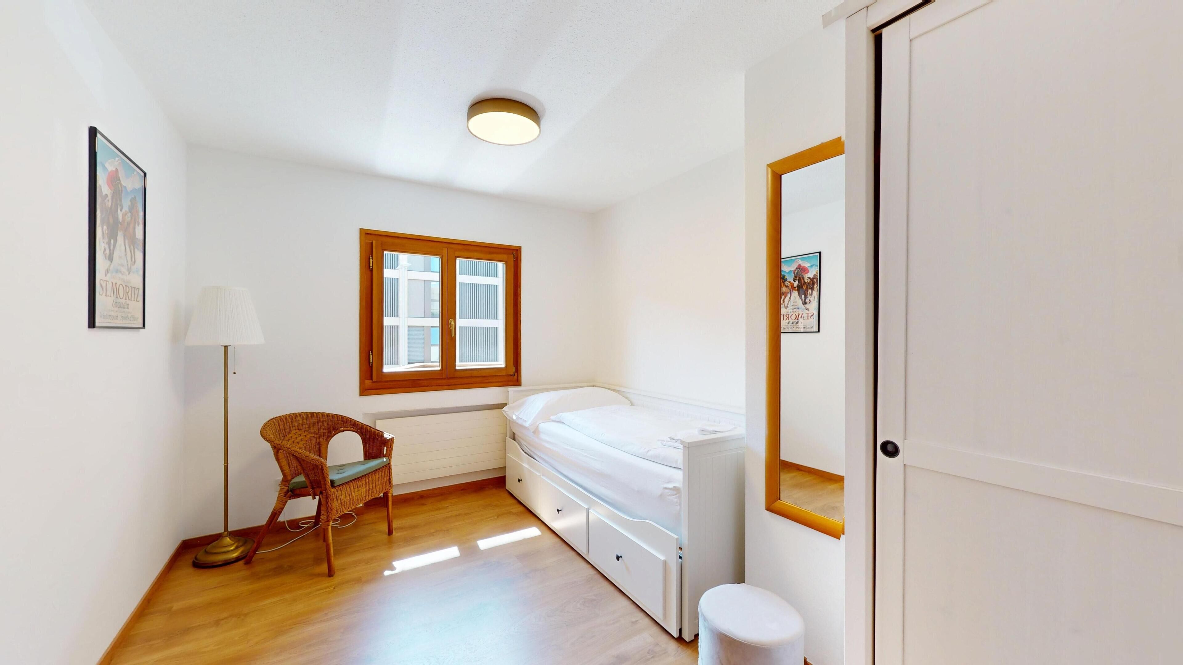A small, clean room with a bed, a chair, a desk, and a window.
