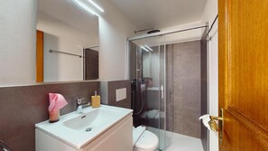 Een moderne badkamer met een wit wastafelmeubel, een spiegel, een douchecabine en een houten deur.