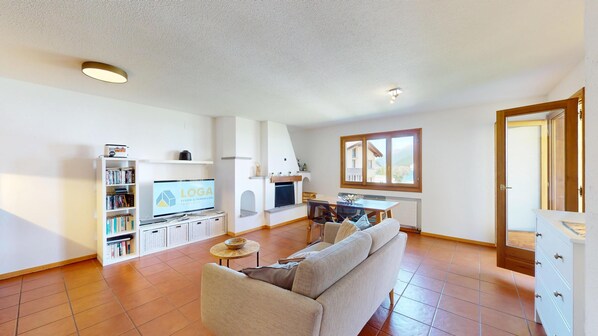 Een woonkamer met een bank, televisie, boekenkast, open haard en een eethoek met een tafel en stoelen.