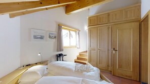 A bedroom with a bed, a desk, a chair, a window with curtains, and wooden cabinets. - 2½-room apartment on the top floor (attic apartment), 64m2. (Silvaplana)